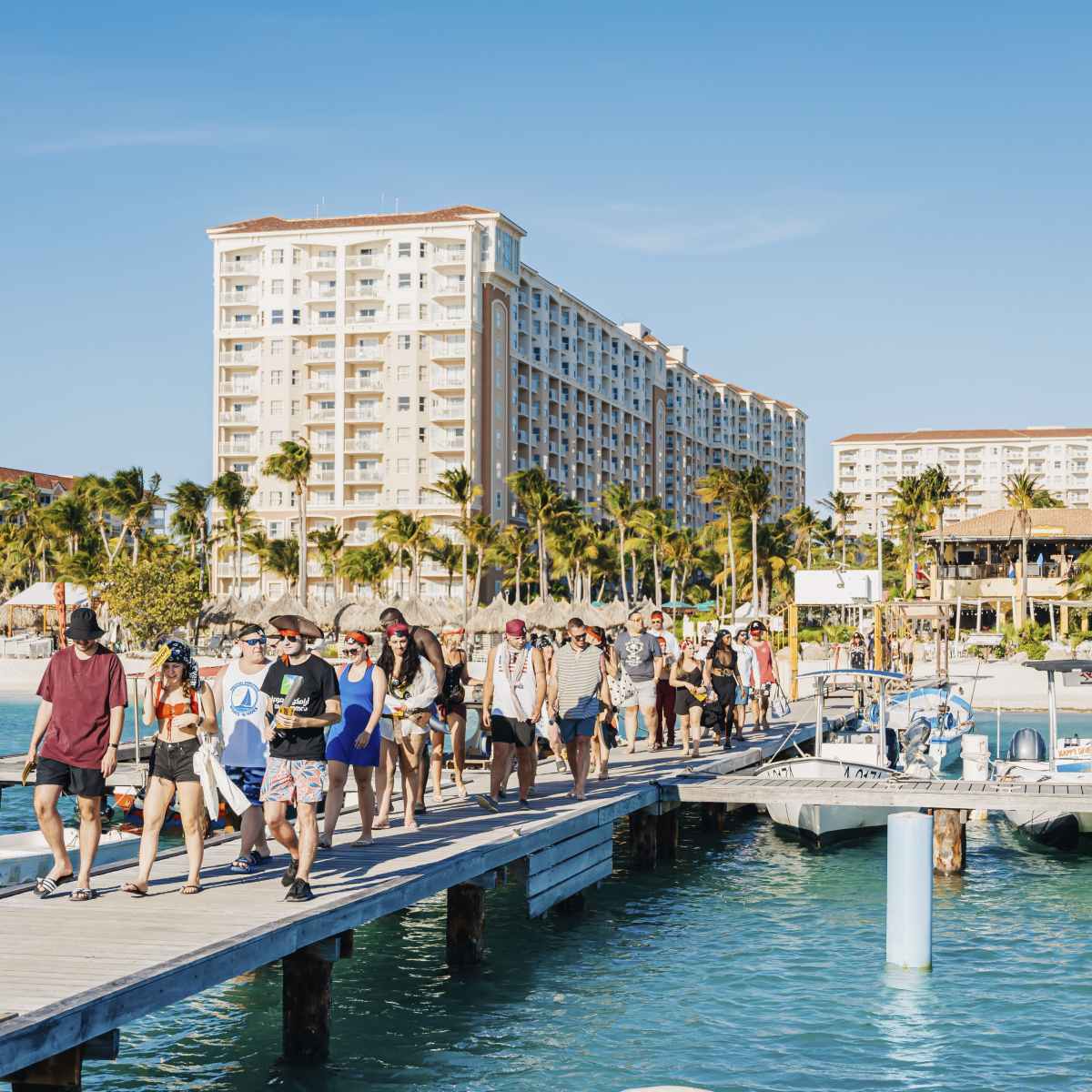 Group of people walking on a pier with tall beachside buildings and palm trees in the background.