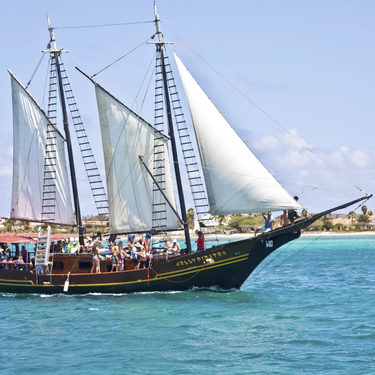 Sailing ship with white sails and passengers, on blue water near a coastline.