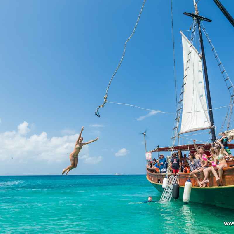 Person swings from a rope off a sailing ship into the turquoise ocean.