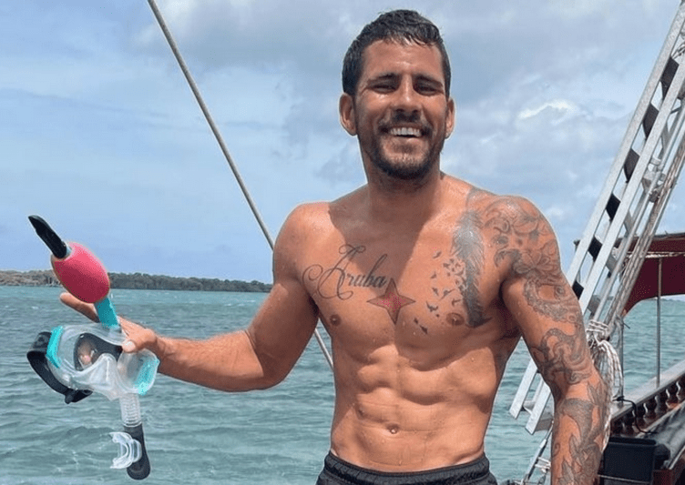 Smiling man on boat holding snorkeling gear, showing tattoos, ocean in background.