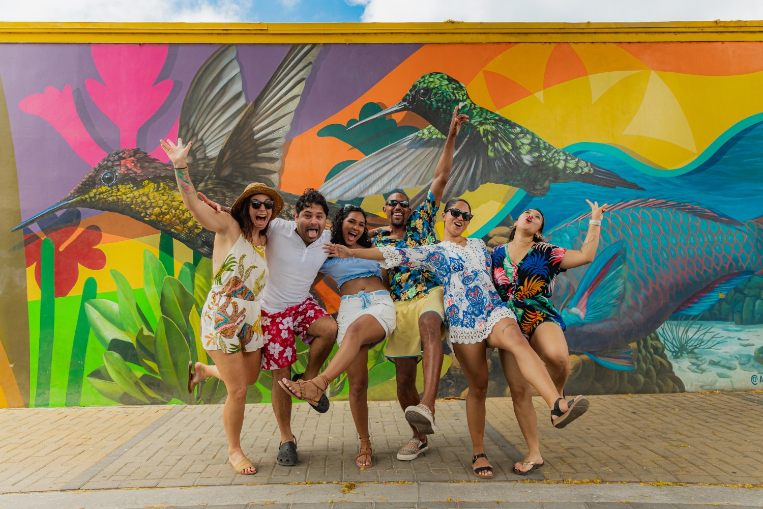 Group of six cheerful people posing in front of a colorful mural of hummingbirds.