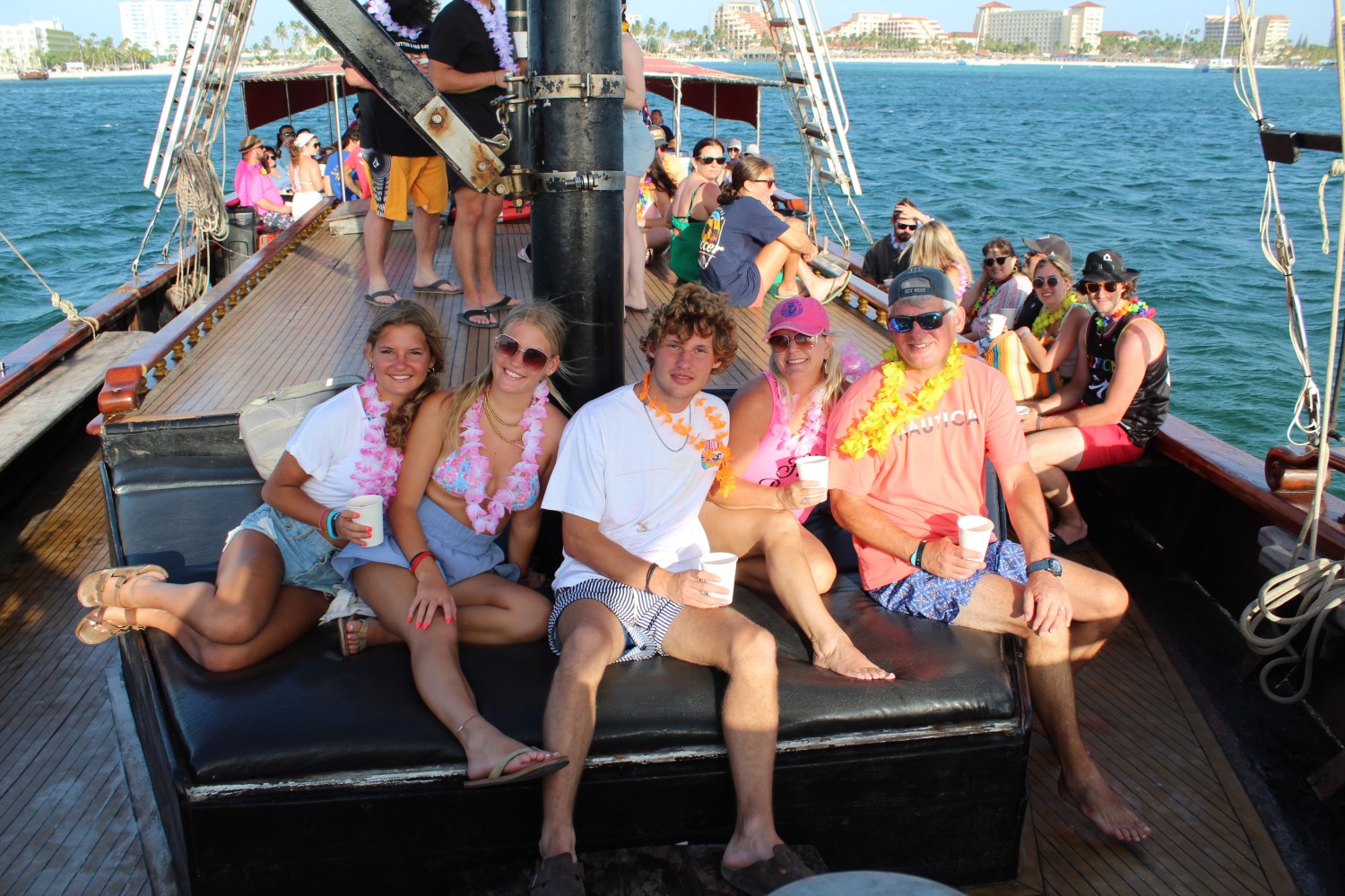 Group of people sitting on a boat deck, wearing leis, with ocean and buildings in the background.