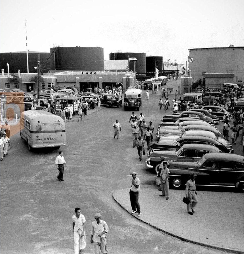 Vintage scene with people, classic cars, and large storage tanks.