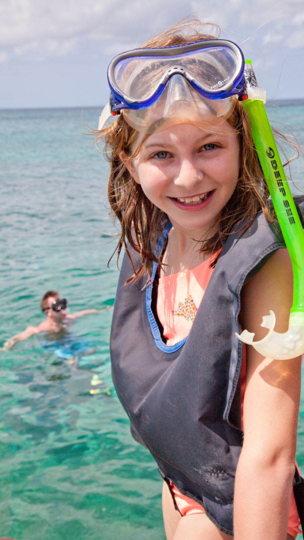 Girl in snorkeling gear smiles on a boat, clear ocean and snorkeler in background.
