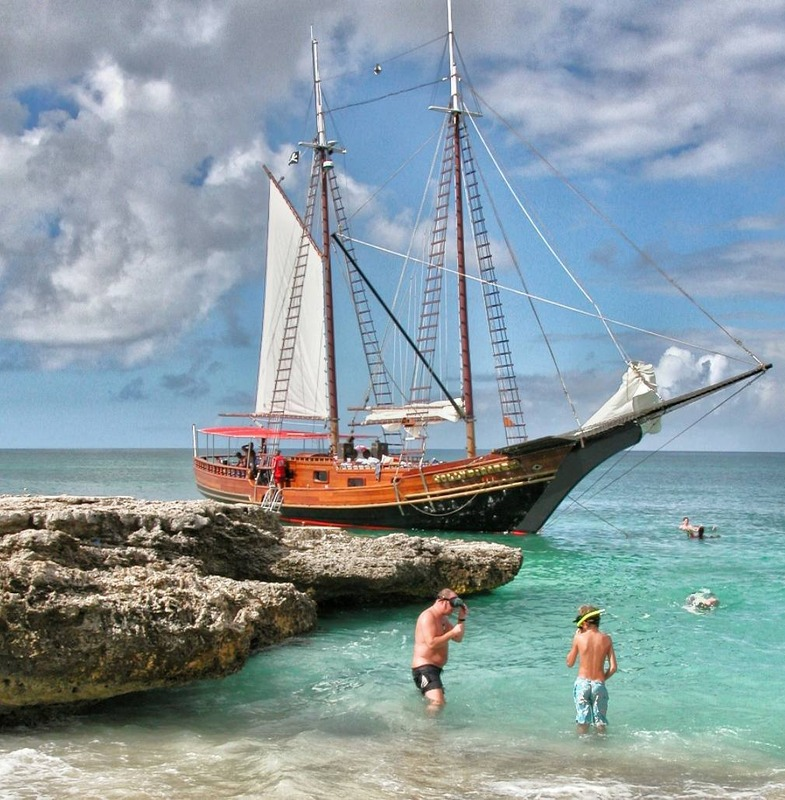 Sailing ship near rocky shore, people swimming in clear water.