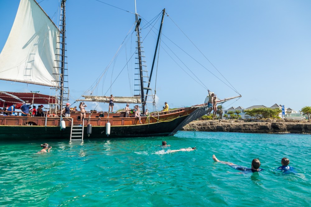 People swimming near a sailing ship in clear turquoise water.