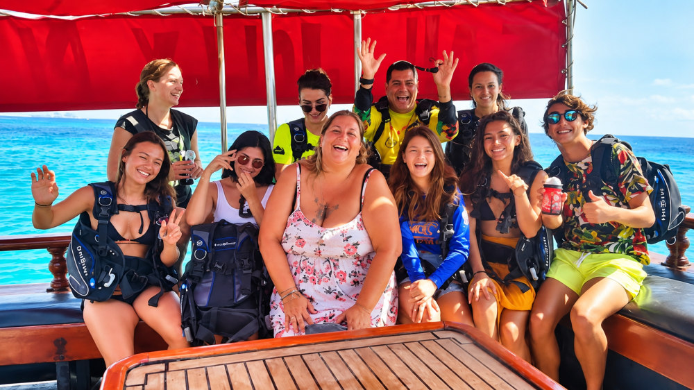 Group of people smiling on a boat with scuba gear, wooden table in foreground.