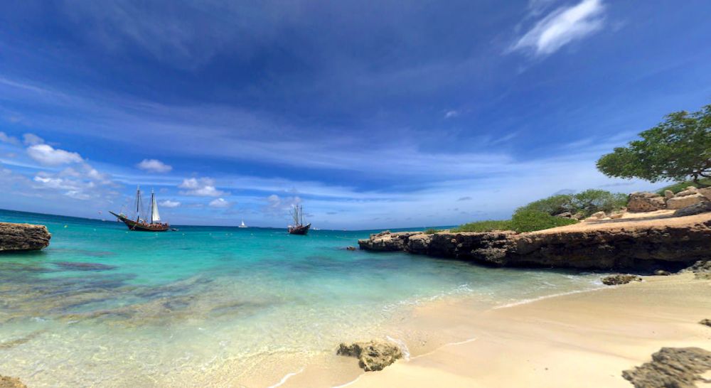 Tropical beach with clear water, rocky shore, and sailboats in the distance under a blue sky.