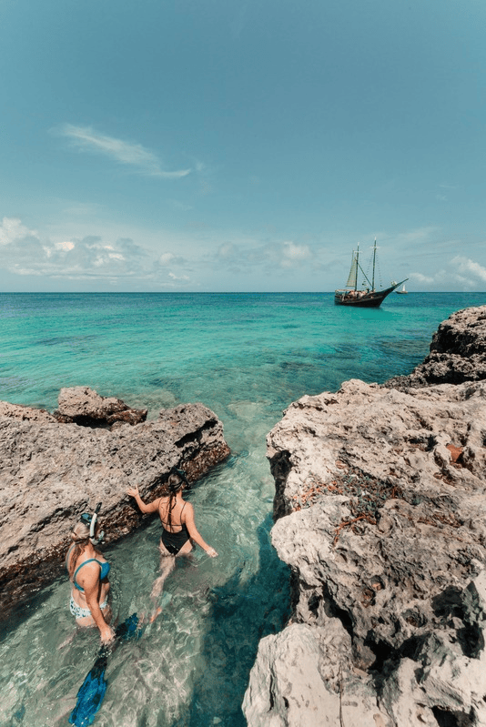 Two snorkelers in clear water near rocky shore, boat in distance.